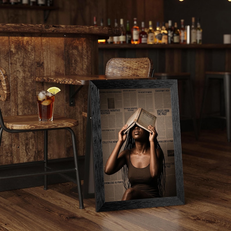May include: A framed vertical print featuring a person with long dark braids wearing a brown tank top, holding an open book over their face. The background consists of a collage of newspaper clippings with text including Voices from the Past.