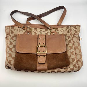 May include: A tan and brown monogram canvas tote bag featuring a front pocket with a dark brown suede lower section, a smooth tan leather flap, and a brass buckle closure. The bag includes two brown leather shoulder straps and brass hardware.