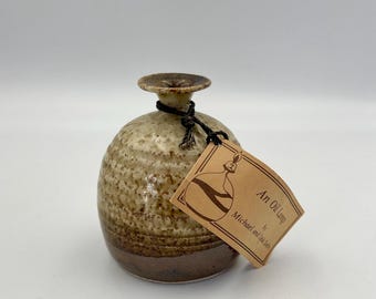 May include: A round ceramic oil lamp with a textured, speckled beige upper body and a smooth, dark brown base. A dark string ties a rectangular tan paper tag to the narrow neck. The tag features a line drawing of the lamp and the text An Oil Lamp by Michael and Lela Seth.