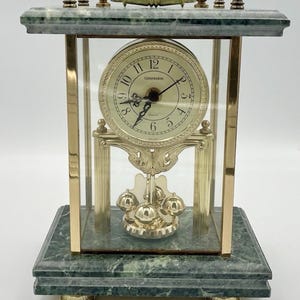 May include: A Commodore quartz mantel clock featuring a green marble base and top, gold-tone metal pillars, and a glass enclosure. The clock face has black numerals and hands, while a rotating pendulum with four gold spheres sits at the bottom.