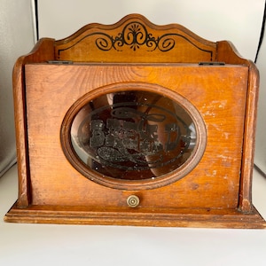 May include: A wooden bread box with a medium brown finish, featuring a hinged front door with an oval glass window. The glass displays the word BREAD in black lettering. The top back panel has a decorative black scrollwork design.