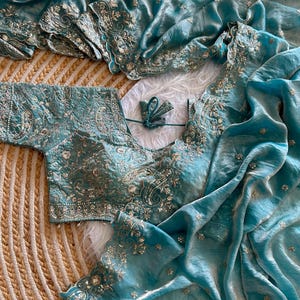 May include: A teal blue saree and matching blouse set featuring intricate silver embroidery and sequin work. The fabric has a subtle sheen, and the borders are finished with a scalloped edge. The blouse includes a tie-up detail at the back.