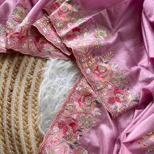 May include: A light pink fabric with a textured, slightly metallic sheen, featuring an intricate scalloped border embroidered with floral motifs in shades of pink, orange, and green, accented with small metallic sequins and beadwork.