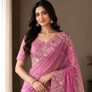May include: A vibrant pink saree featuring intricate floral embroidery along the borders and bodice. The fabric has a subtle, textured sheen, and the design includes delicate pink and white flower motifs that create a cohesive, detailed pattern.