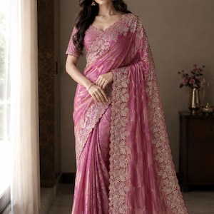 May include: A vibrant pink saree featuring intricate gold embroidery along the scalloped borders and a matching short-sleeved blouse. The fabric has a subtle sheen with a textured pattern, draped elegantly with detailed floral motifs throughout.