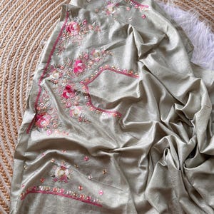 May include: A sage green fabric piece featuring intricate floral embroidery in shades of pink, white, and gold. The embroidery includes detailed rose motifs and decorative borders with pink stitching, creating a textured and layered appearance.