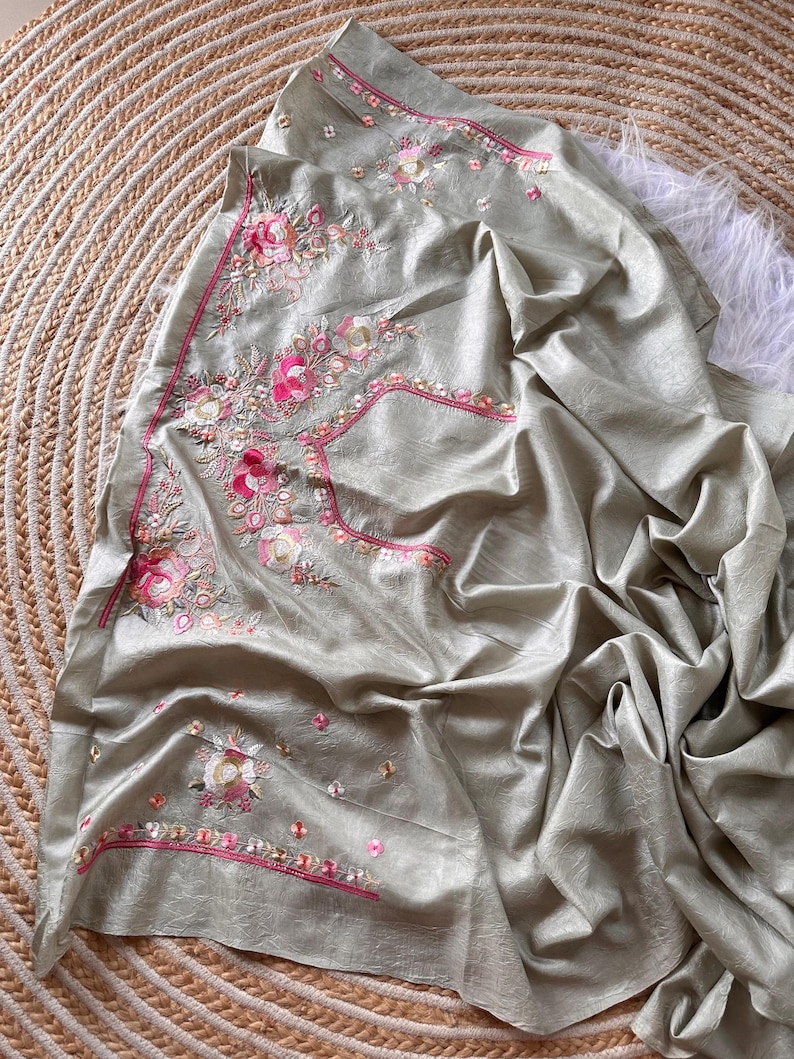 May include: A sage green fabric piece featuring intricate floral embroidery in shades of pink, white, and gold. The embroidery includes detailed rose motifs and decorative borders with pink stitching, creating a textured and layered appearance.