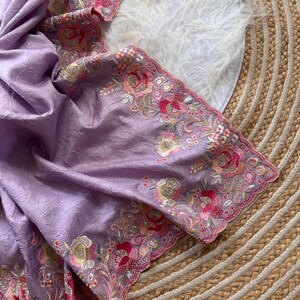 May include: A light purple fabric with a crinkled texture featuring an intricate scalloped border. The edge is decorated with detailed floral embroidery in shades of pink, orange, and gold, accented with small sparkling sequins throughout.