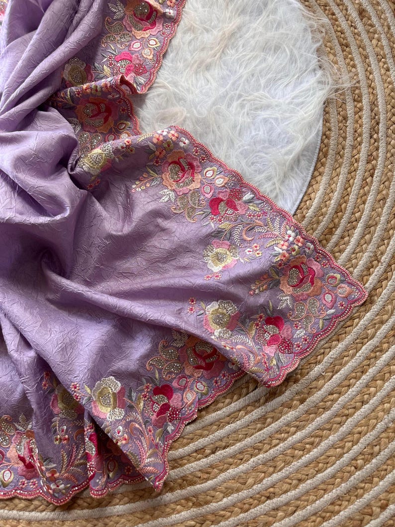 May include: A light purple fabric with a crinkled texture featuring an intricate scalloped border. The edge is decorated with detailed floral embroidery in shades of pink, orange, and gold, accented with small sparkling sequins throughout.