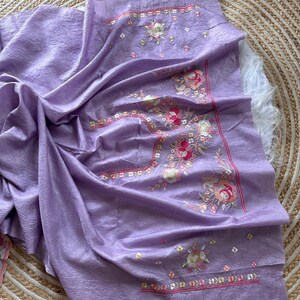 May include: A lavender-colored fabric piece featuring a textured surface and intricate floral embroidery. The design includes pink, white, and green threadwork with small metallic accents arranged in a decorative pattern along the edges.