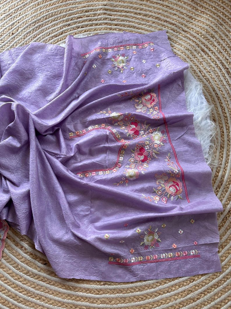 May include: A lavender-colored fabric piece featuring a textured surface and intricate floral embroidery. The design includes pink, white, and green threadwork with small metallic accents arranged in a decorative pattern along the edges.