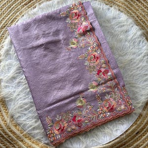 May include: A lavender fabric with a crinkled texture featuring an intricate floral border. The embroidery showcases roses in shades of pink, orange, and green, accented with small metallic beads and sequins along a scalloped, finished edge.