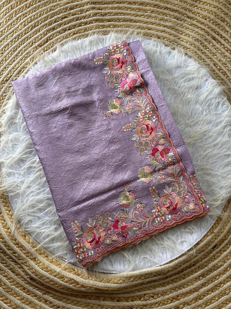 May include: A lavender fabric with a crinkled texture featuring an intricate floral border. The embroidery showcases roses in shades of pink, orange, and green, accented with small metallic beads and sequins along a scalloped, finished edge.