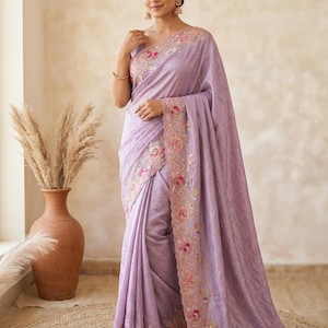 May include: A lavender-colored saree featuring intricate floral embroidery along the borders. The fabric has a subtle sheen and a textured appearance, with pink and orange floral motifs accented by delicate beadwork throughout the edges.