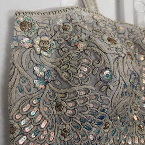 May include: A light grey crop top with detailed embroidery and sequin embellishments. The top features floral and feather-like patterns in silver, gold, and iridescent sequins. The straps are also embellished.