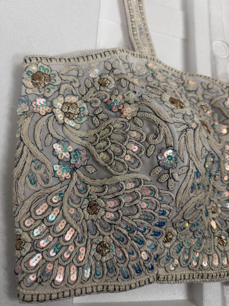 May include: A light grey crop top with detailed embroidery and sequin embellishments. The top features floral and feather-like patterns in silver, gold, and iridescent sequins. The straps are also embellished.
