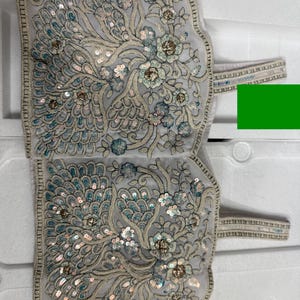 May include: A light grey textile with detailed floral embroidery and sequin embellishments. The design features stylised flowers and peacock-like feather patterns in teal, silver, and gold. The edges are trimmed with a delicate border and straps.