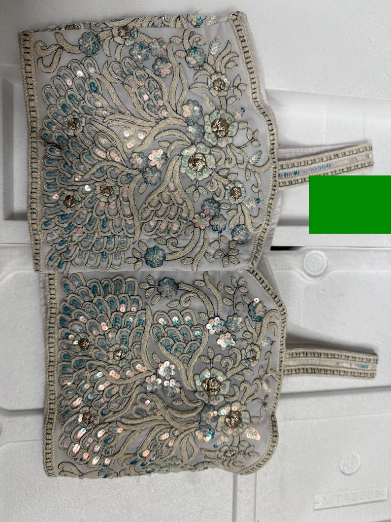 May include: A light grey textile with detailed floral embroidery and sequin embellishments. The design features stylised flowers and peacock-like feather patterns in teal, silver, and gold. The edges are trimmed with a delicate border and straps.