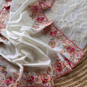 May include: A white fabric piece featuring a textured surface and a scalloped edge. The border is decorated with intricate floral embroidery in shades of pink, orange, and green, accented with gold-toned metallic thread and small clear beads.