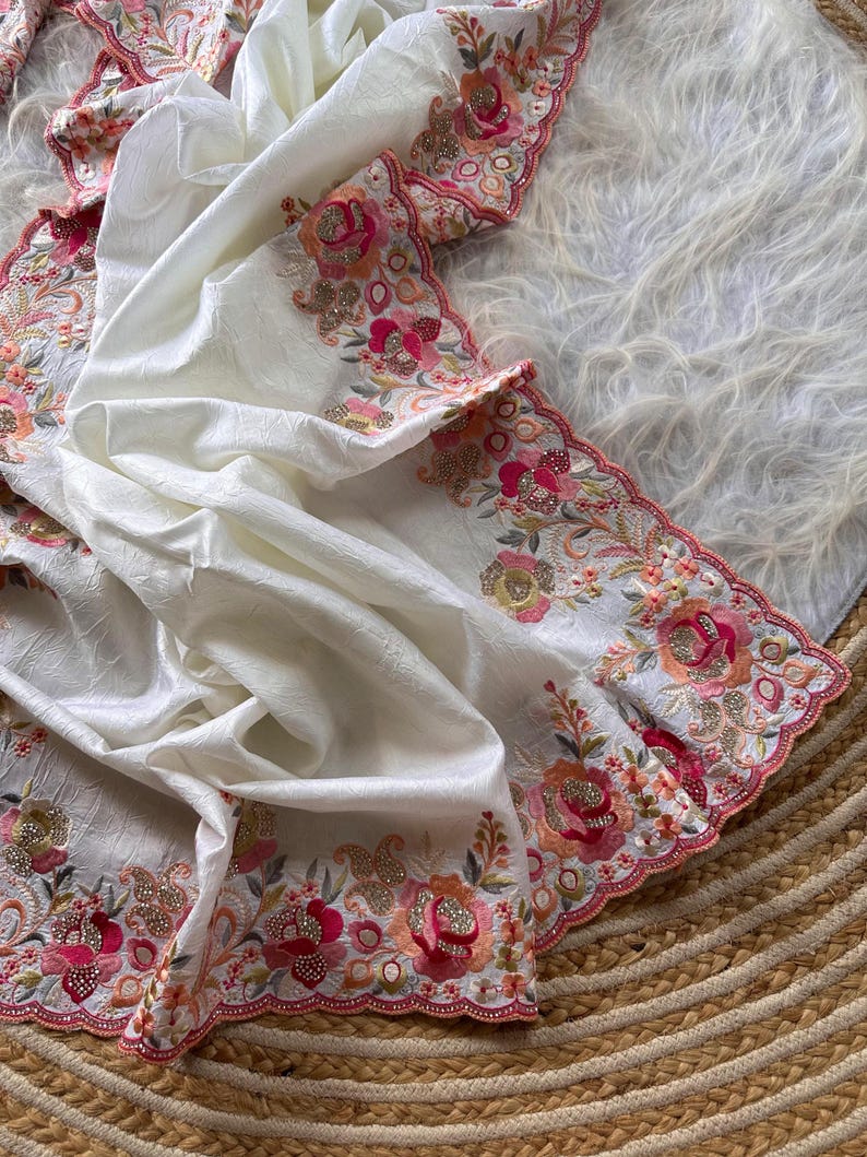 May include: A white fabric piece featuring a textured surface and a scalloped edge. The border is decorated with intricate floral embroidery in shades of pink, orange, and green, accented with gold-toned metallic thread and small clear beads.