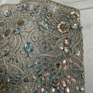 May include: A light grey, intricately embroidered top featuring floral and peacock feather patterns. The top is adorned with sequins in shades of blue, pink, and gold. The edges are trimmed with a beaded border, creating a detailed and elegant appearance.