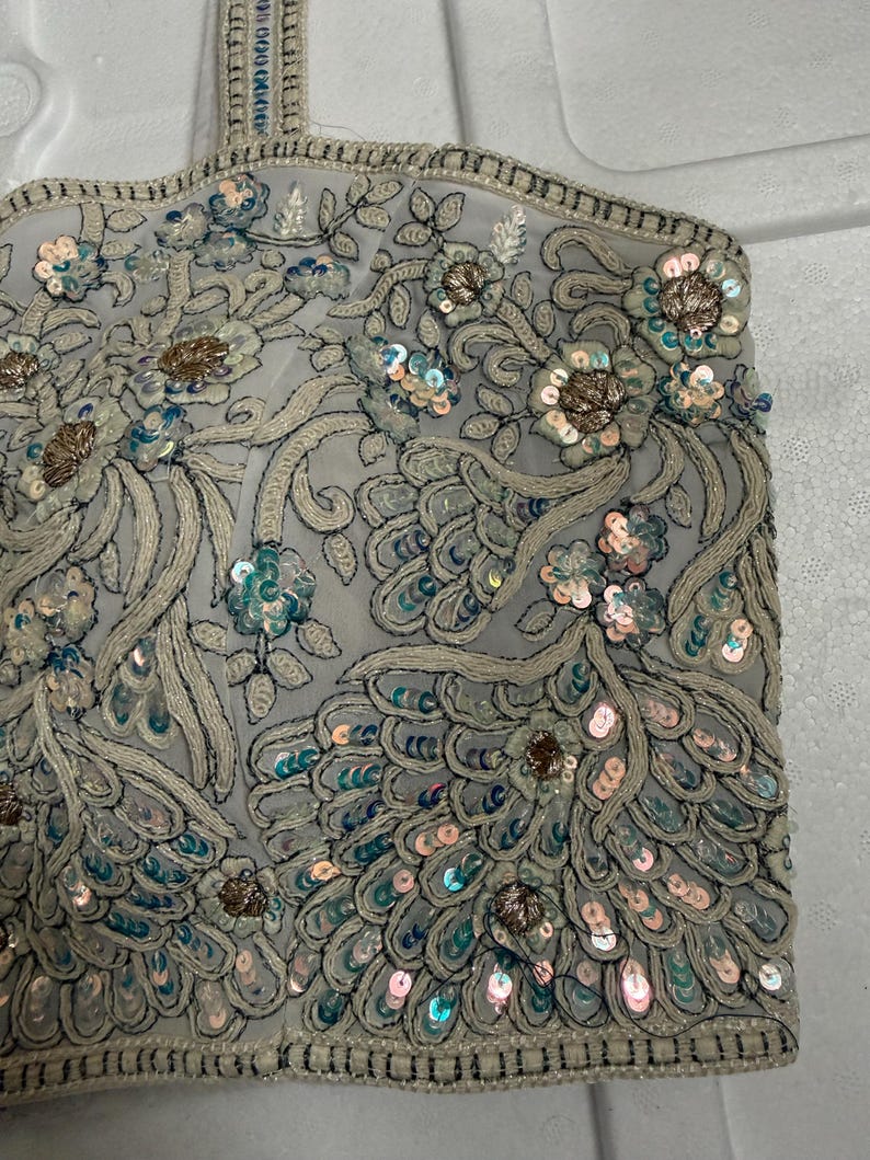 May include: A light grey, intricately embroidered top featuring floral and peacock feather patterns. The top is adorned with sequins in shades of blue, pink, and gold. The edges are trimmed with a beaded border, creating a detailed and elegant appearance.
