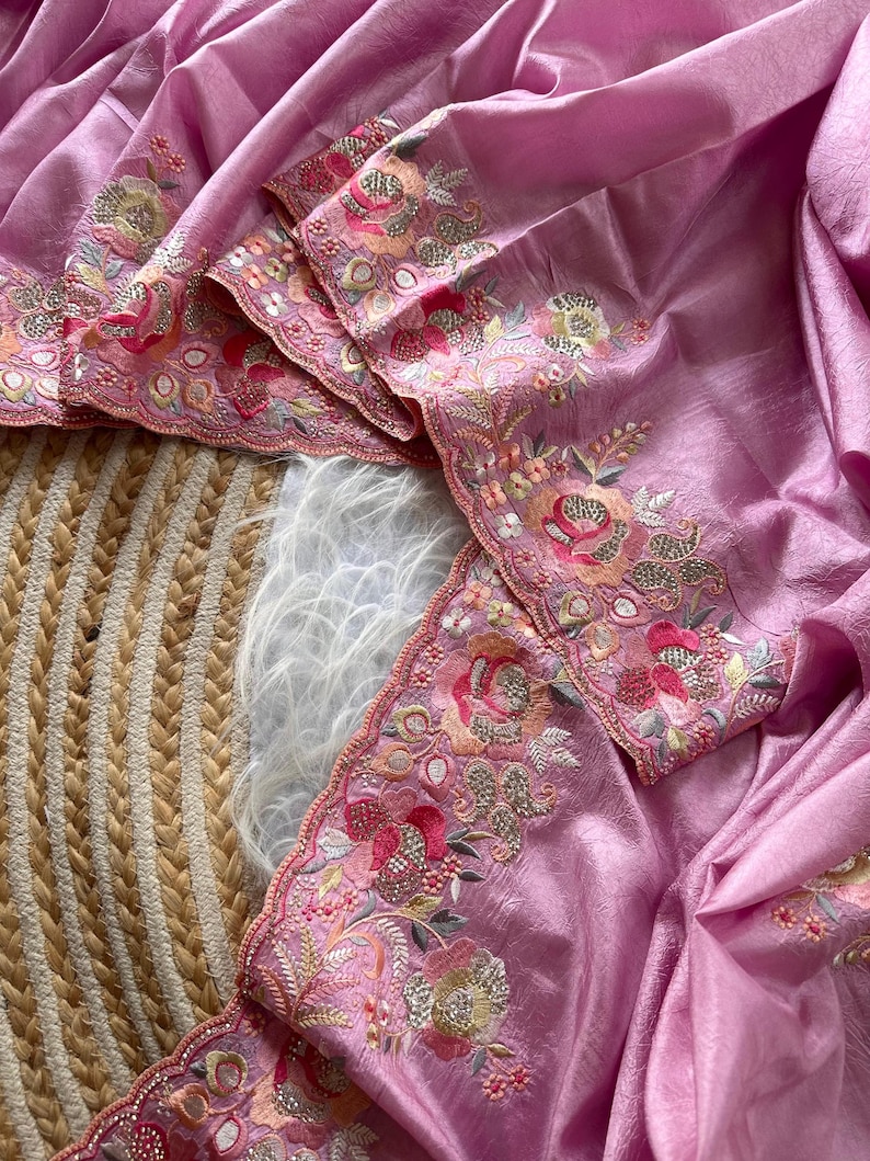 May include: A light pink fabric with a textured, slightly metallic sheen, featuring an intricate scalloped border embroidered with floral motifs in shades of pink, orange, and green, accented with small metallic sequins and beadwork.