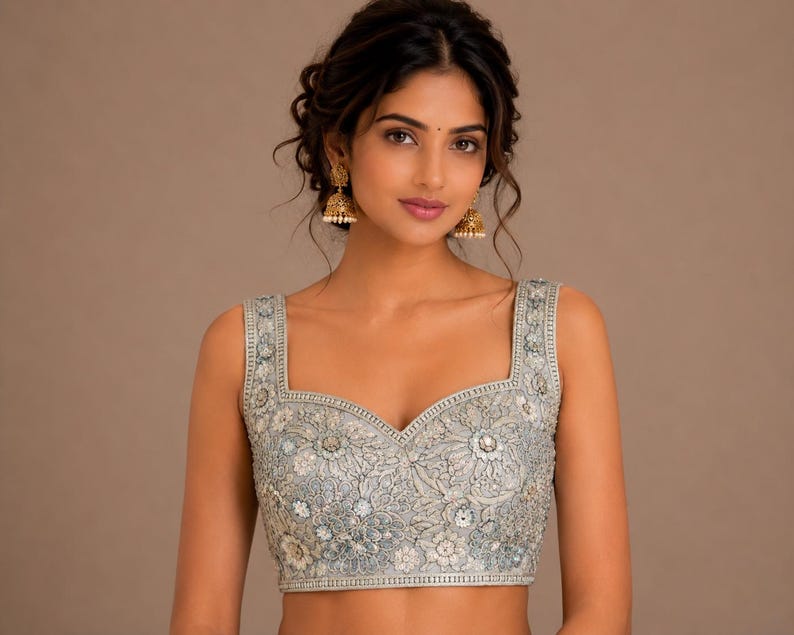 May include: A pale blue embroidered sleeveless top with a square neckline. The top has detailed floral patterns and embellishments. The model is wearing gold earrings. The background is a plain brown colour.