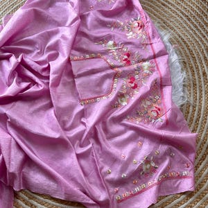 May include: A light pink fabric garment featuring intricate floral embroidery in shades of pink, white, and green. The design includes detailed needlework patterns with small sequins and beads scattered throughout the floral motifs on the smooth material.