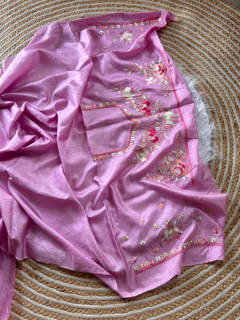 May include: A light pink fabric garment featuring intricate floral embroidery in shades of pink, white, and green. The design includes detailed needlework patterns with small sequins and beads scattered throughout the floral motifs on the smooth material.