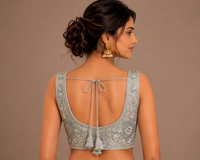 May include: A silver sleeveless blouse with detailed floral embroidery and embellishments. The back has a tie closure with tassels. The blouse is a formal wear garment.