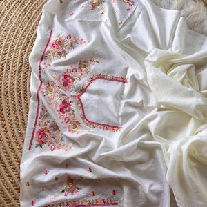May include: A white fabric garment featuring intricate floral embroidery in shades of pink, orange, and gold. The embroidery includes detailed flower motifs and decorative borders with metallic accents, set against a textured, off-white material.