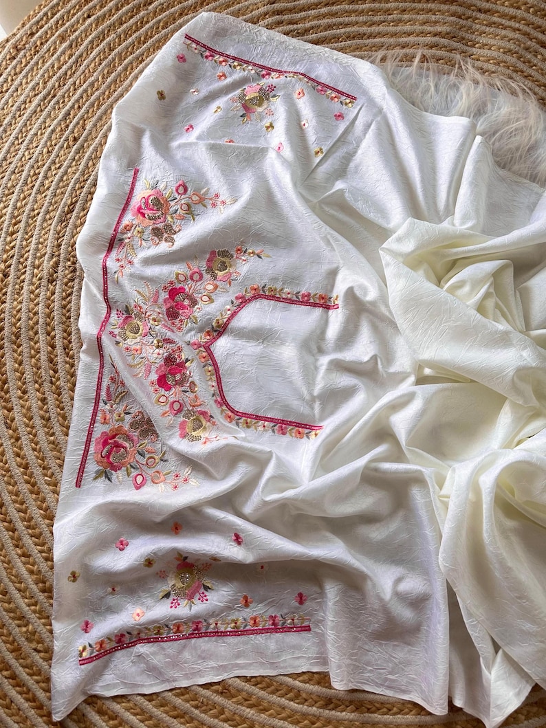 May include: A white fabric garment featuring intricate floral embroidery in shades of pink, orange, and gold. The embroidery includes detailed flower motifs and decorative borders with metallic accents, set against a textured, off-white material.