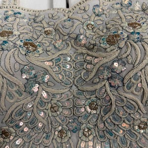 May include: Close-up of a pale grey top with detailed embroidery and embellishments. The design includes floral patterns, sequins, and beads in silver, blue, and gold. The straps are also embellished.