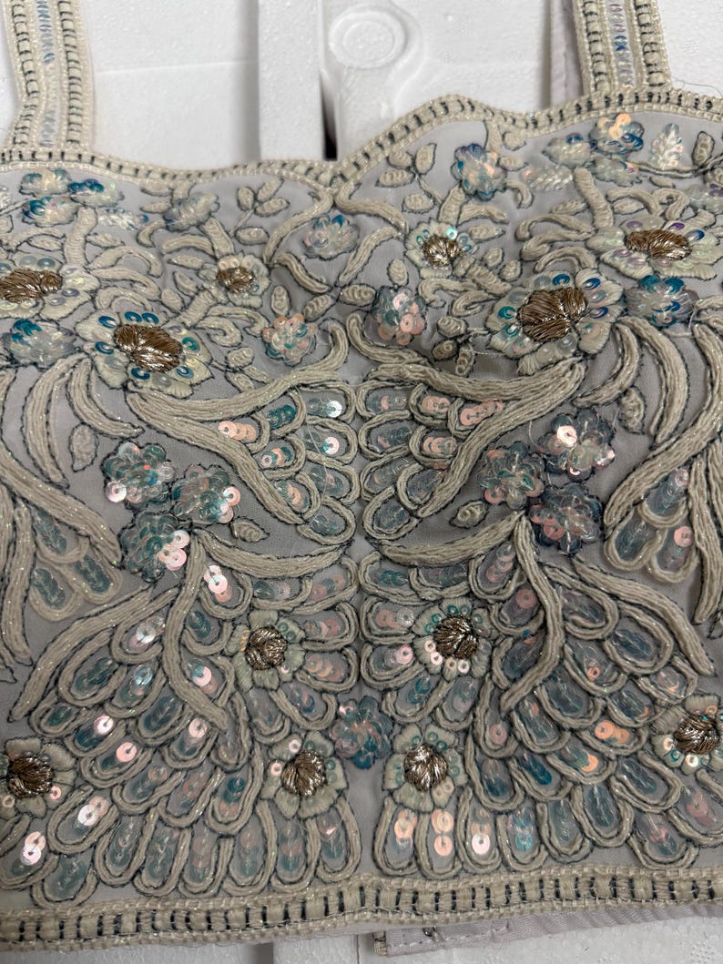May include: Close-up of a pale grey top with detailed embroidery and embellishments. The design includes floral patterns, sequins, and beads in silver, blue, and gold. The straps are also embellished.
