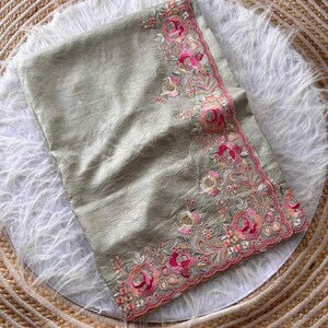May include: A sage green fabric with a crinkled texture featuring an ornate border of embroidered pink and peach roses, small white flowers, and green leaves. The edge of the fabric is finished with a scalloped trim and intricate beadwork.