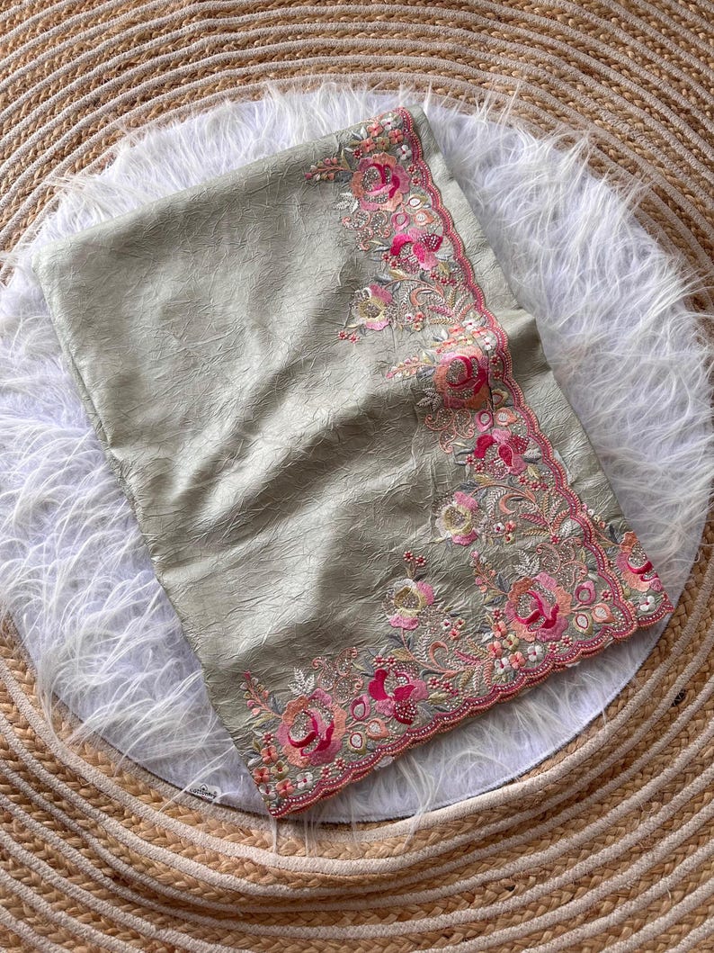 May include: A sage green fabric with a crinkled texture featuring an ornate border of embroidered pink and peach roses, small white flowers, and green leaves. The edge of the fabric is finished with a scalloped trim and intricate beadwork.