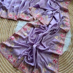 May include: A lavender fabric textile featuring a scalloped edge with intricate floral embroidery. The embroidery displays a vibrant color palette of pink, orange, green, and gold threads, creating detailed rose and leaf patterns along the border.