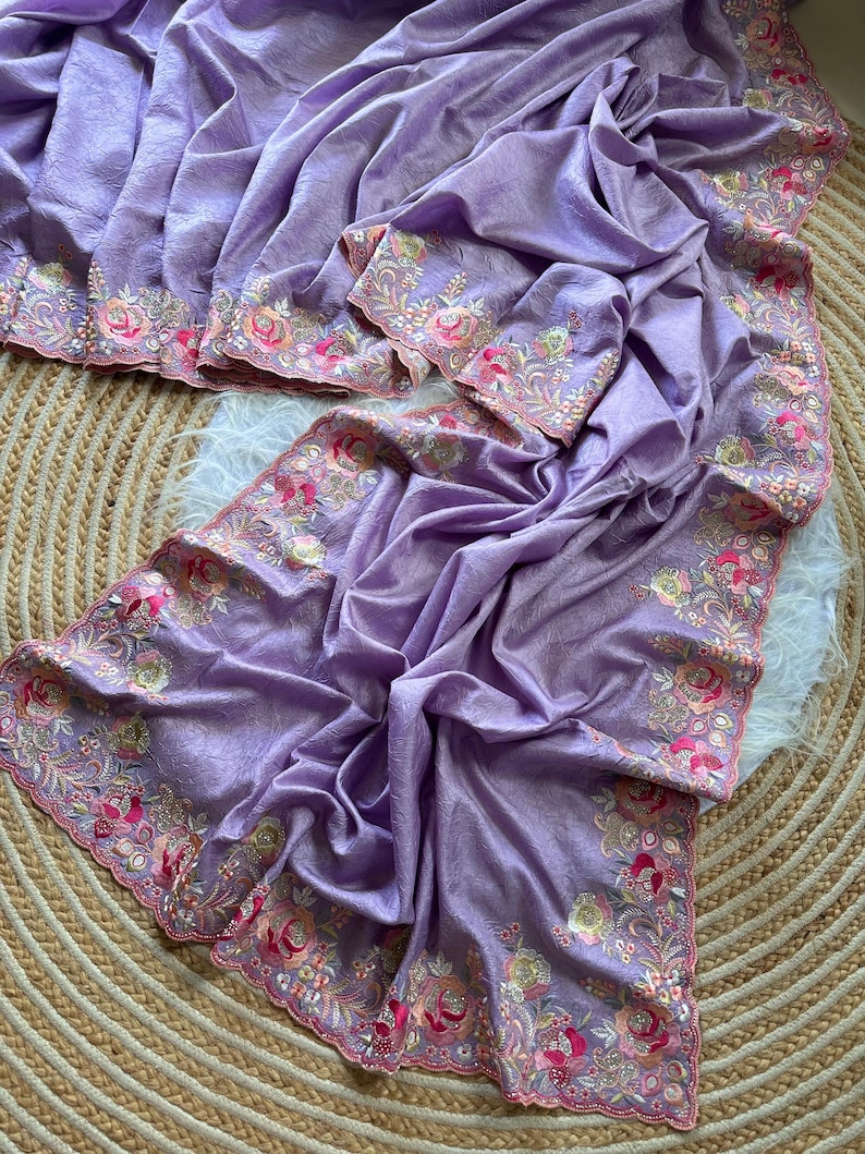 May include: A lavender fabric textile featuring a scalloped edge with intricate floral embroidery. The embroidery displays a vibrant color palette of pink, orange, green, and gold threads, creating detailed rose and leaf patterns along the border.