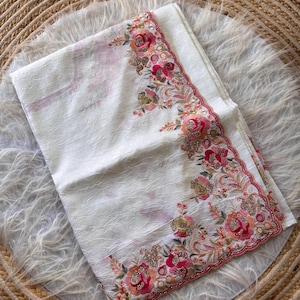 May include: A white fabric piece with a textured, crinkled surface. The edge features an intricate floral embroidery pattern in shades of pink, orange, and gold, accented with small sparkling sequins and a scalloped, stitched border finish.