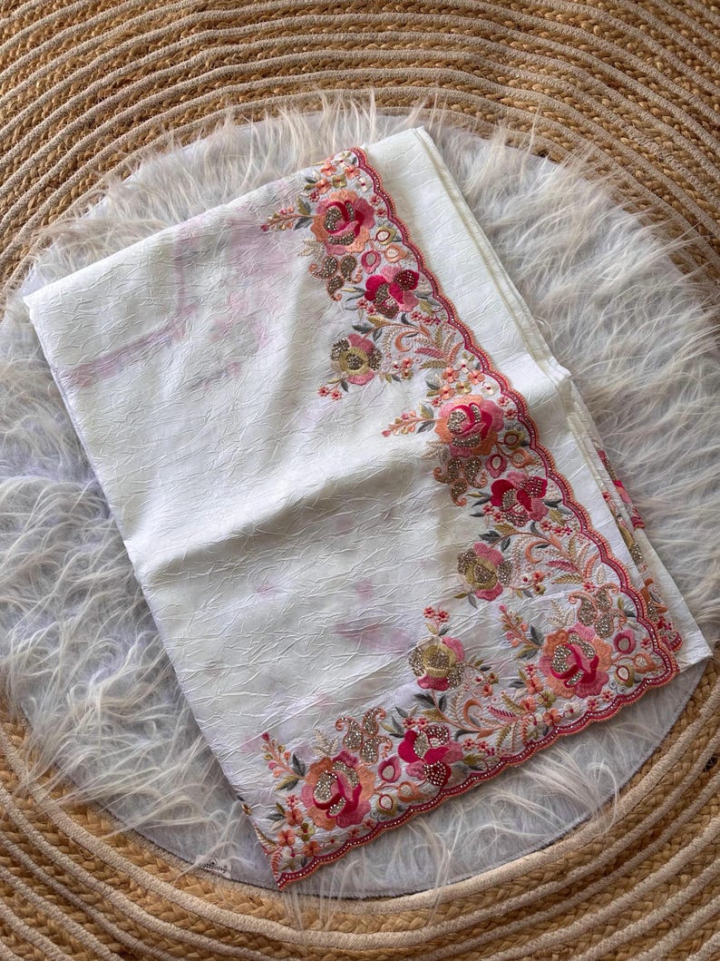 May include: A white fabric piece with a textured, crinkled surface. The edge features an intricate floral embroidery pattern in shades of pink, orange, and gold, accented with small sparkling sequins and a scalloped, stitched border finish.