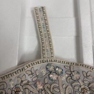 May include: Close-up of a light grey top with floral embroidery and sequin embellishments. The top features a delicate design with white, gold, and teal accents. The strap is adorned with small, shimmering sequins.
