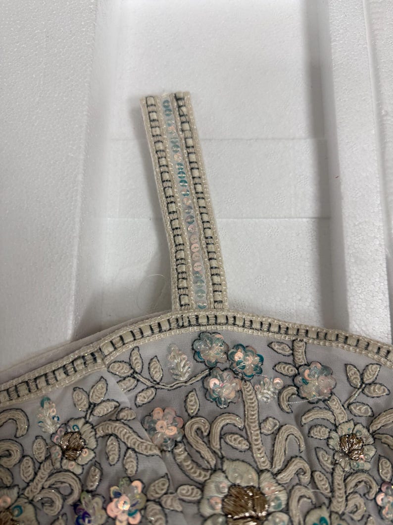 May include: Close-up of a light grey top with floral embroidery and sequin embellishments. The top features a delicate design with white, gold, and teal accents. The strap is adorned with small, shimmering sequins.