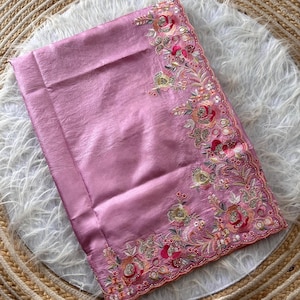 May include: A folded mauve fabric textile featuring an intricate embroidered border along one edge. The embroidery displays floral motifs in shades of pink, gold, and green, accented with small reflective sequins and metallic thread detailing.