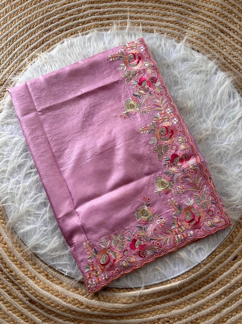 May include: A folded mauve fabric textile featuring an intricate embroidered border along one edge. The embroidery displays floral motifs in shades of pink, gold, and green, accented with small reflective sequins and metallic thread detailing.
