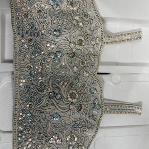 May include: A light grey, intricately embroidered garment with floral and feather-like patterns. The embroidery features silver and iridescent sequins, with straps on each side. The design is detailed and ornate, suggesting a formal or decorative purpose.
