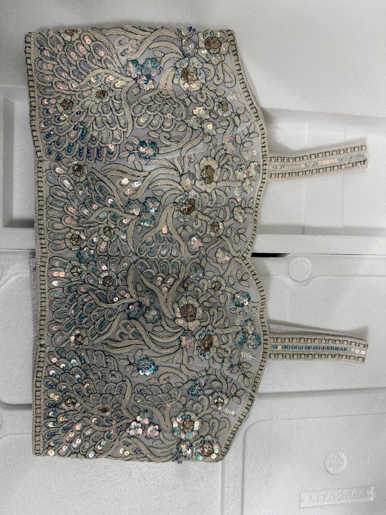 May include: A light grey, intricately embroidered garment with floral and feather-like patterns. The embroidery features silver and iridescent sequins, with straps on each side. The design is detailed and ornate, suggesting a formal or decorative purpose.