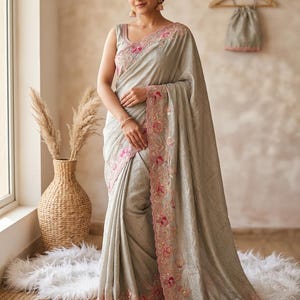 May include: A light gray sleeveless saree featuring a scalloped border with intricate pink and gold floral embroidery. The fabric has a subtle sheen, and the matching blouse is displayed on a wooden hanger in the background.