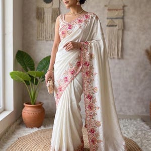 May include: A cream-colored saree featuring intricate floral embroidery in shades of pink, peach, and green along the borders and bodice. The garment includes a sleeveless blouse and a draped fabric panel with scalloped edges and detailed stitching.
