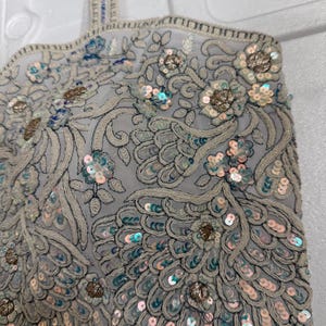 May include: A light grey, intricately embroidered top with floral and feather-like patterns. The design features sequins in shades of silver, blue, and gold. The edges are finished with a beaded trim, creating a detailed and elegant look.